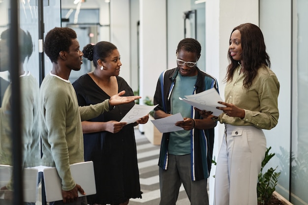 Grupo de jovens empresários negros de pé no corredor do escritório moderno e discutindo gráficos e ...