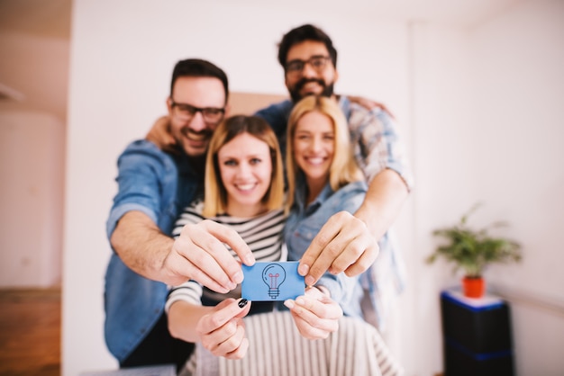 Grupo de jovens encantadores felizes segurando juntos um pequeno papel azul com uma lâmpada para idéias criativas e inovadoras