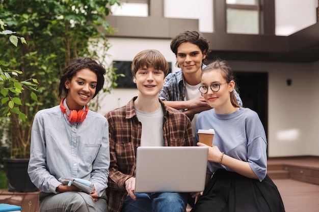 Grupo de jovens estudantes alegres, sentados no banco e trabalhando juntos no laptop, enquanto felizes