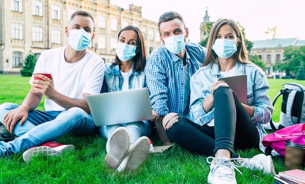 Grupo de jovens estudantes com máscaras médicas com laptop e livros estão estudando juntos na Universidade. Amigos sentados na grama ao ar livre.