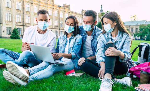 Grupo de jovens estudantes com máscaras médicas com laptop e livros estão estudando juntos na Universidade. Amigos sentados na grama ao ar livre.