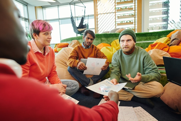 Grupo de jovens gerentes financeiros interculturais em trajes casuais discutindo papéis com diagramas e gráficos em reunião de trabalho no chão