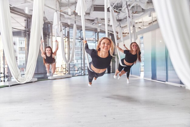 Grupo de jovens mulheres esportivas estendendo-se em redes aéreas de ioga na academia com janelas panorâmicas