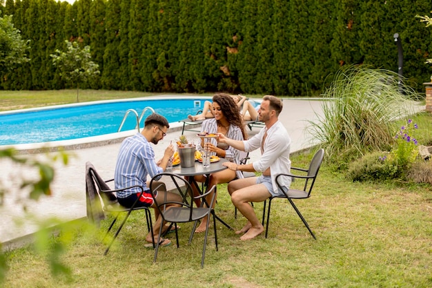 Grupo de jovens torcendo com bebidas e comendo frutas à beira da piscina no jardim