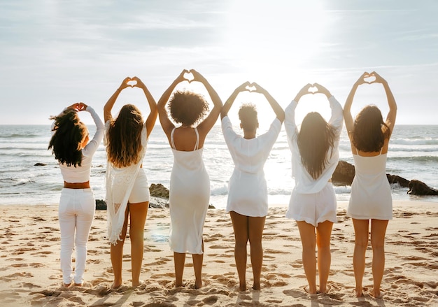 Grupo de mulheres jovens desfrutando do pôr-do-sol no mar fazendo símbolo de lareira com as mãos em linha