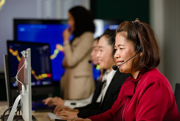 Grupo de operadores de atendimento ao cliente feminino asiáticos usa fones de ouvido de microfone sentar sorrindo na frente gráfico diário gráfico computador monitor tela falando e negociando comprar vender mercado de ações on-line com o cliente.