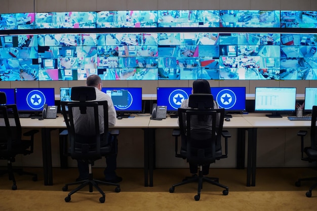 Grupo de operadores de data center de segurança trabalhando em uma sala de monitoramento CCTV olhando para vários monitores Oficiais monitorando várias telas para atividades suspeitas Equipe trabalhando no System Contr