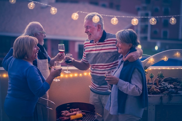 Grupo de pessoas alegres e felizes amigos idosos tilintando e desfrutando juntos do jantar ao ar livre no terraço de casa com vista para a cidade