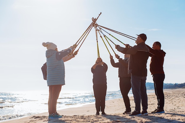 Grupo de pessoas fazendo caminhada nórdica cruzando bastões de trekking juntos no ar contra as ondas do mar na praia fria