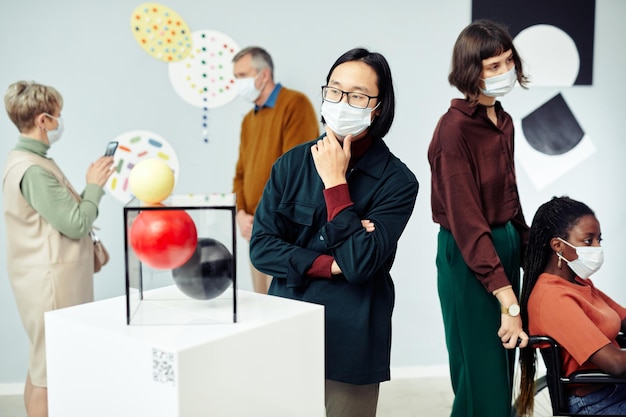 Grupo de pessoas multiétnicas modernas usando máscaras protetoras olhando objetos de arte contemporânea em exposição na galeria