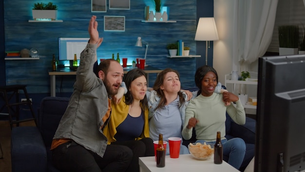 Grupo de pessoas multirraciais torcendo pelo time de futebol enquanto está sentado no sofá à noite na sala de estar. Amigos multiétnicos passando um tempo juntos assistindo a um jogo de futebol na televisão