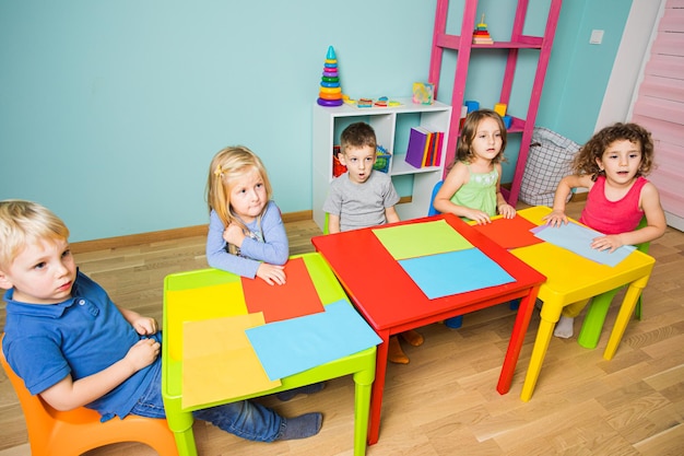 Grupo de pré-escolares está sentado em uma mesa no jardim de infância As crianças estão se preparando para criar um cartão de papel colorido