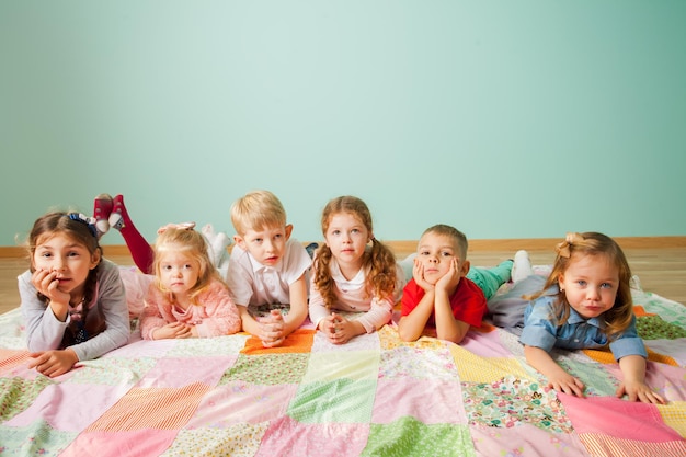 Grupo de seis crianças bonitinhas, meninos e meninas de diferentes idades, deitado no chão no cobertor de retalhos colorido em casa na sala de estar olhando para a câmera com interesse