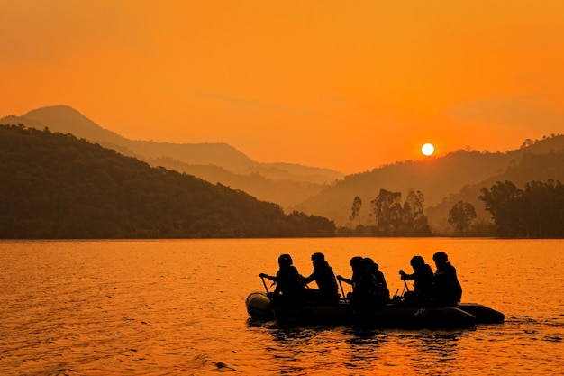 Grupo de soldados ajuda a barco a remo para mirar no crepúsculo, montanha e sol no fundo.