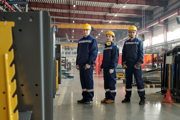 Grupo de trabalhadores confiantes da fábrica de compressores, vestindo capacetes amarelos e roupas de trabalho azuis, em fila na oficina da fábrica