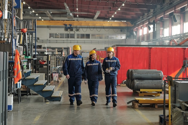 Grupo de trabalhadores ou engenheiros da fábrica contemporânea movendo-se ao longo de uma grande oficina com equipamentos industriais e conversando