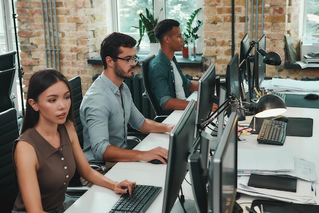 Grupo de trabalho em equipe de jovens funcionários trabalhando em computadores enquanto estão sentados em espaço aberto moderno