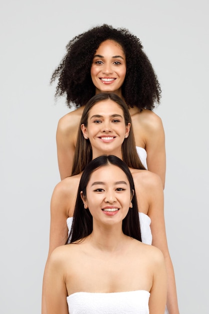 Grupo de três mulheres bonitas e diversas sorrindo para a câmera em pé e posando sobre estúdio cinza
