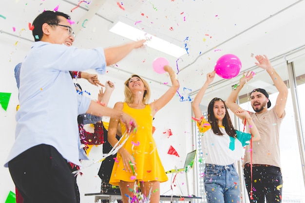 Grupo de disfrutar de los jóvenes celebrando lanzando confeti mientras anima y salta en la fiesta en la sala blanca.