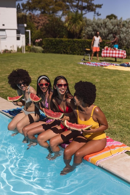 Grupo diversificado de amigas comendo melancia sentada à beira da piscina conversando. Sair e relaxar ao ar livre no verão.