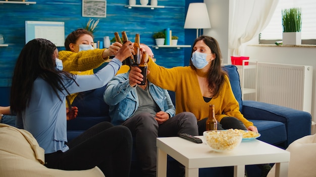 Grupo diversificado de amigos assistindo TV juntos comédia de comédia comendo lanches e bebendo cerveja, tilintando garrafas e se divertindo em uma nova festa normal, usando máscara facial de proteção contra o vírus covid 19