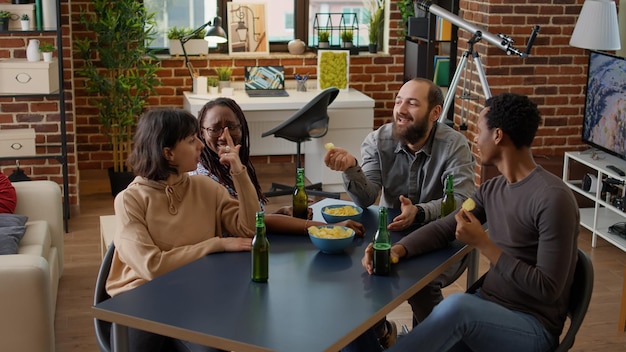 Grupo diversificado de amigos conversando e se divertindo na sala de estar, bebendo cerveja e comendo batatas fritas. Pessoas felizes conversando na reunião de atividades de lazer, curtindo a discussão juntos.