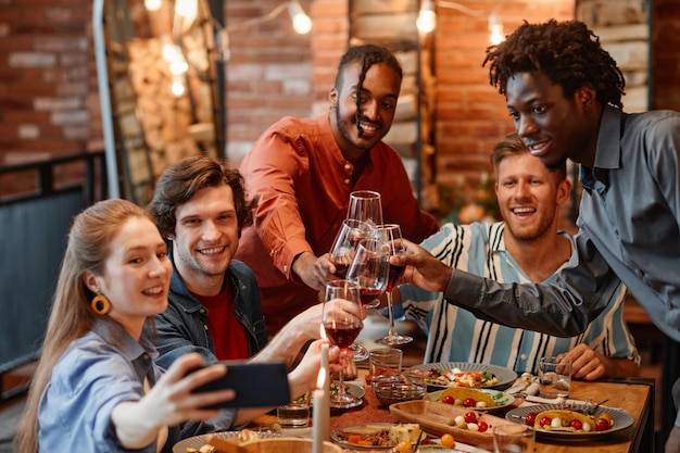 Grupo diversificado de amigos tirando foto de selfie na mesa durante o jantar e copos tinindo