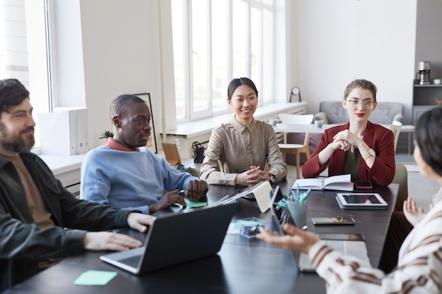 Grupo diversificado de empresários à mesa durante reunião de briefing no escritório, foco na mulher asiática sorridente, compartilhando ideias