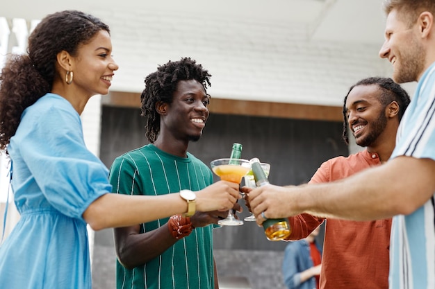 Grupo diversificado de jovens desfrutando de bebidas na festa no terraço ao ar livre e brindando com copos