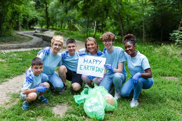 Grupo diversificado de pessoas recolhendo lixo no parque voluntário serviço comunitário voluntários internacionais felizes segurando cartaz com mensagem de 'feliz dia da terra'