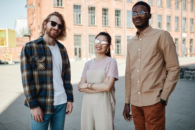 Grupo diversificado de três jovens olhando para a câmera em pé ao ar livre na cidade, todos usando óculos escuros