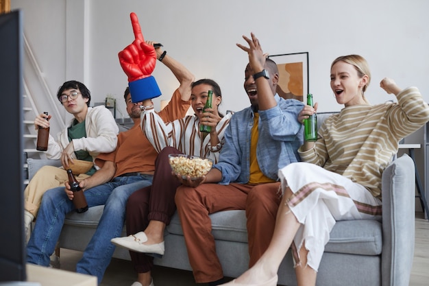 Grupo diverso de fanáticos de los deportes viendo el partido en casa y animando alegremente levantando las manos