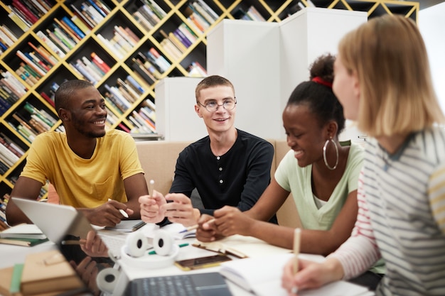 Grupo diverso de jóvenes que estudian juntos en la mesa en la biblioteca de la universidad se centran en sonrientes m ...