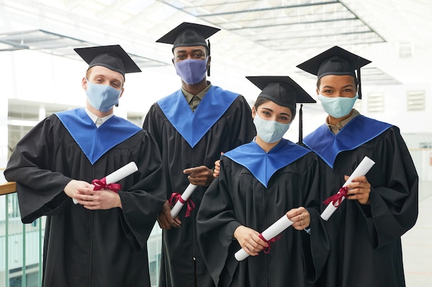 Grupo diverso de jóvenes con togas y máscaras de graduación mientras posan en interiores en el interior de la universidad