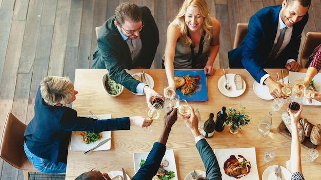 Un grupo diverso de profesionales, incluidos hombres y mujeres de varias etnias, disfrutan de una cena de celebración y brindis. La mesa está llena de comida y bebidas que destacan una reunión alegre.