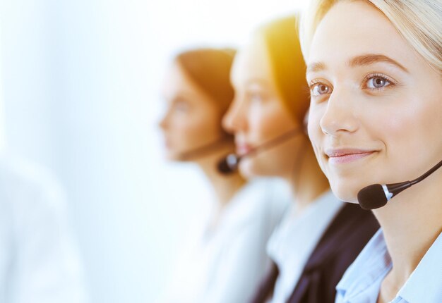 Grupo de diversos operadores telefónicos trabajando en una oficina soleada. Hermosa mujer de negocios con auriculares consultando a clientes. Centro de llamadas y concepto de gente de negocios.