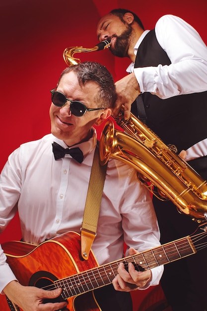 Foto grupo de dos músicos, banda masculina de jazz, guitarrista y saxofonista con trajes clásicos improvisan con instrumentos musicales en un estudio de fondo rojo