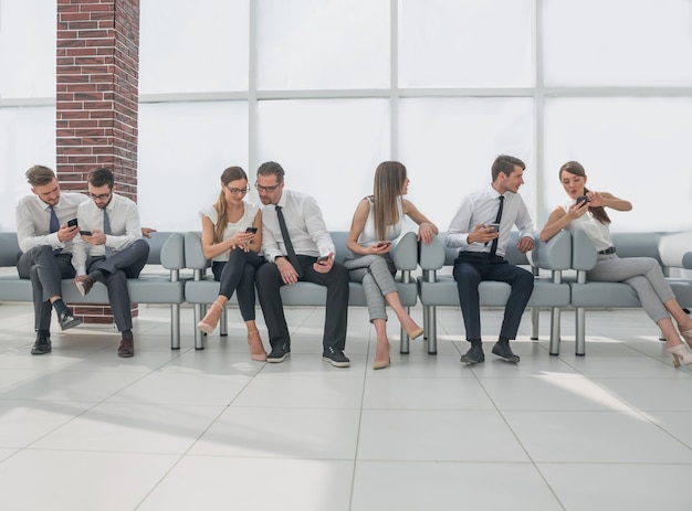 Un grupo de empleados usa teléfonos inteligentes mientras están sentados en la foto del vestíbulo de la oficina con espacio para copiar