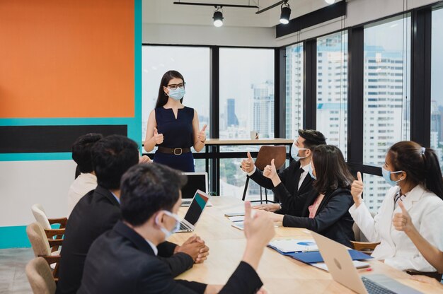 Grupo empresarial multiétnico usando máscara facial e mostrando o polegar para cima parabenizado com uma executiva sobre o acordo de cooperação da empresa durante a reunião no novo escritório normal