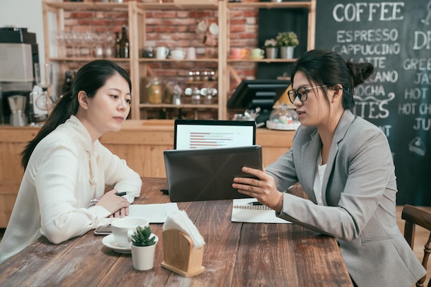 Grupo de empresarios reunidos en una cafetería. Empresaria asiática en traje apuntando a la computadora de la tableta que muestra el catálogo de productos para los inversores de los clientes. dos señoras de la oficina hablando sobre el teclado digital del proyecto