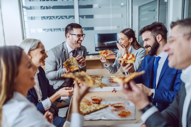 Grupo de empresarios sentados en la sala de juntas, charlando, riendo y comiendo pizza para el almuerzo.