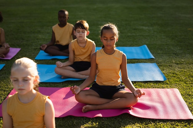 Grupo de estudiantes diversos practicando yoga y meditando sentados en colchonetas de yoga en el jardín de la escuela