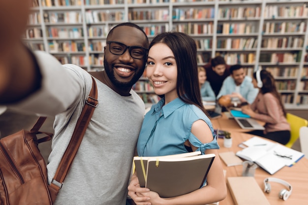 Grupo de estudiantes étnicos multiculturales en la biblioteca. Chico negro y chica asiática tomando selfie en teléfono.