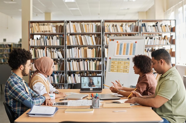 Grupo de estudiantes extranjeros que tienen una lección en línea con un maestro mientras están sentados en la biblioteca