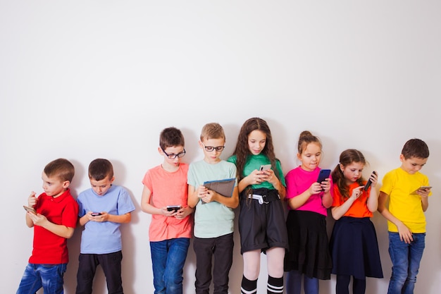 Grupo de estudiantes de primaria felices con teléfonos inteligentes sobre pared blanca Tecnología de nueva generación y concepto de amistad