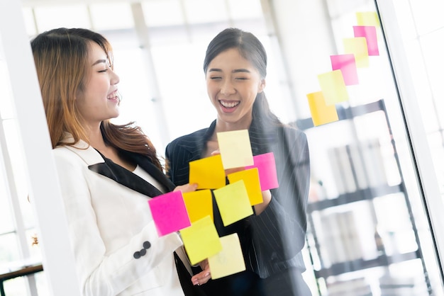 Grupo de exitosas mujeres empresarias asiáticas trabajan en equipo Reunión de lluvia de ideas con notas de papel adhesivas de colores en la pared de vidrio para nuevas ideas Uso de metodología ágil para negocios en una oficina de inicio tecnológico