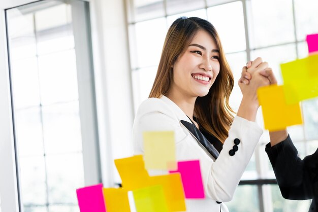 Grupo de exitosas mujeres empresarias asiáticas trabajan en equipo Reunión de lluvia de ideas con notas de papel adhesivas de colores en la pared de vidrio para nuevas ideas Uso de metodología ágil para negocios en una oficina de inicio tecnológico