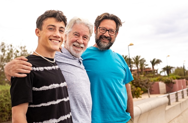 Grupo familiar multigeracional em pé ao ar livre à beira-mar, abraçando uns aos outros e sorrindo. avô, filho e neto adolescente.