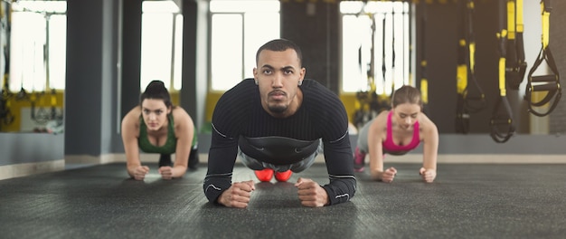 Grupo de fitness haciendo entrenamiento de ejercicios de plancha con entrenador en el interior. Hombres y mujeres jóvenes haciendo ejercicio en el gimnasio. Estilo de vida saludable, concepto de gimnasia, espacio de copia