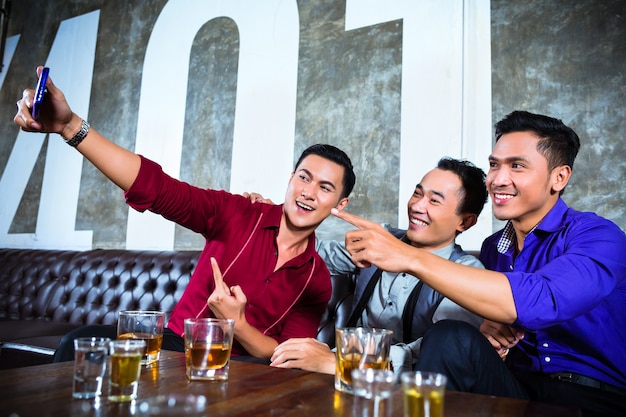 Grupo de gente de fiesta asiática de jóvenes amigos tomando fotos o selfies con su teléfono móvil o celular en un elegante club nocturno